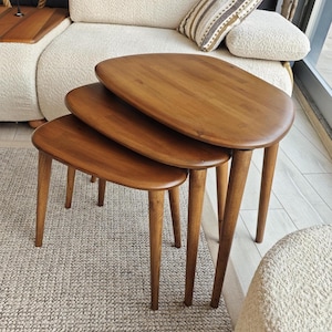 May include: A set of three nesting tables crafted from medium-toned wood, each featuring a smooth, rounded, oval-shaped tabletop and four tapered legs. The tables are arranged in a cascading formation, with each table decreasing in size.