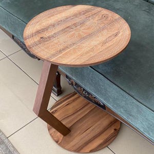 Puede incluir: Una mesa auxiliar redonda de madera con un acabado de veta natural en tonos marrones. El tablero presenta un intrincado patrón floral circular grabado con láser y un borde tipo cuerda. Tiene una pata inclinada unida a una base circular.
