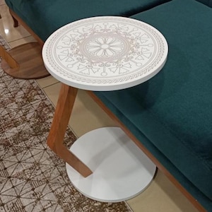 Puede incluir: Una mesa auxiliar redonda con una superficie blanca que presenta un patrón geométrico detallado de estilo mandala en tonos claros. La mesa tiene una sola pata de madera inclinada conectada a una base circular blanca y sólida.