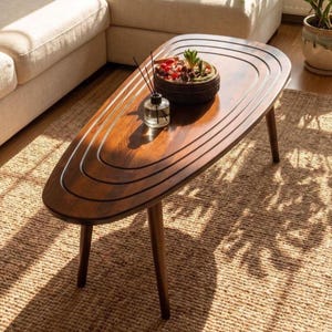 May include: A dark brown wooden coffee table with an elongated oval shape and concentric carved grooves on the surface. The table features three tapered legs and holds a small glass reed diffuser and a decorative bowl filled with colorful stones.