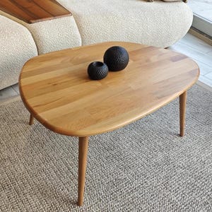May include: A light brown wooden coffee table with a smooth, rounded, organic-shaped top and three tapered legs. Two small, round, black decorative objects with a textured, dimpled surface are placed on the center of the wooden tabletop.