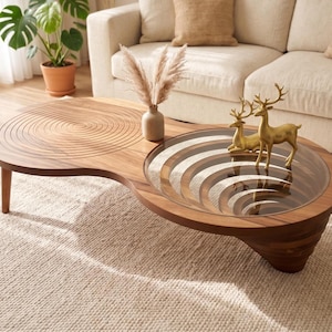 May include: A kidney-shaped wooden coffee table featuring a carved concentric circle pattern on one side and a circular glass inset with a layered wood design underneath on the other. Two gold-colored deer figurines sit on the glass surface.