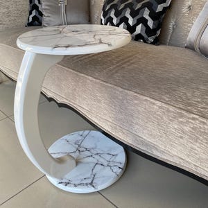 May include: A C-shaped side table featuring a round top and a matching round base, both finished in a white faux marble pattern with dark grey veining. The table has a smooth, glossy white curved support column connecting the top and base.