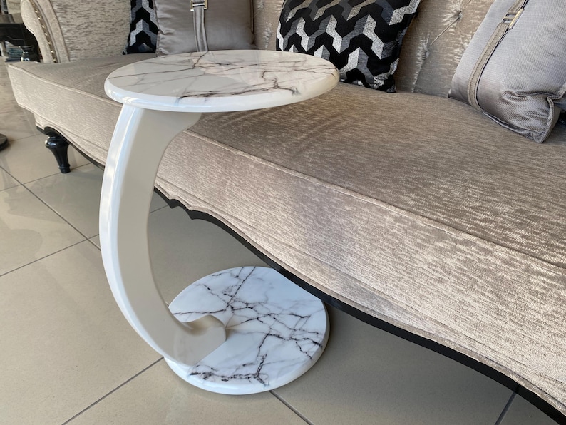 May include: A C-shaped side table featuring a round top and a matching round base, both finished in a white faux marble pattern with dark grey veining. The table has a smooth, glossy white curved support column connecting the top and base.