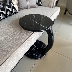 May include: A C-shaped side table featuring a circular black faux marble top and base with white veining. The table has a curved, glossy black support column that connects the top and base, designed to slide over the edge of a sofa or chair.