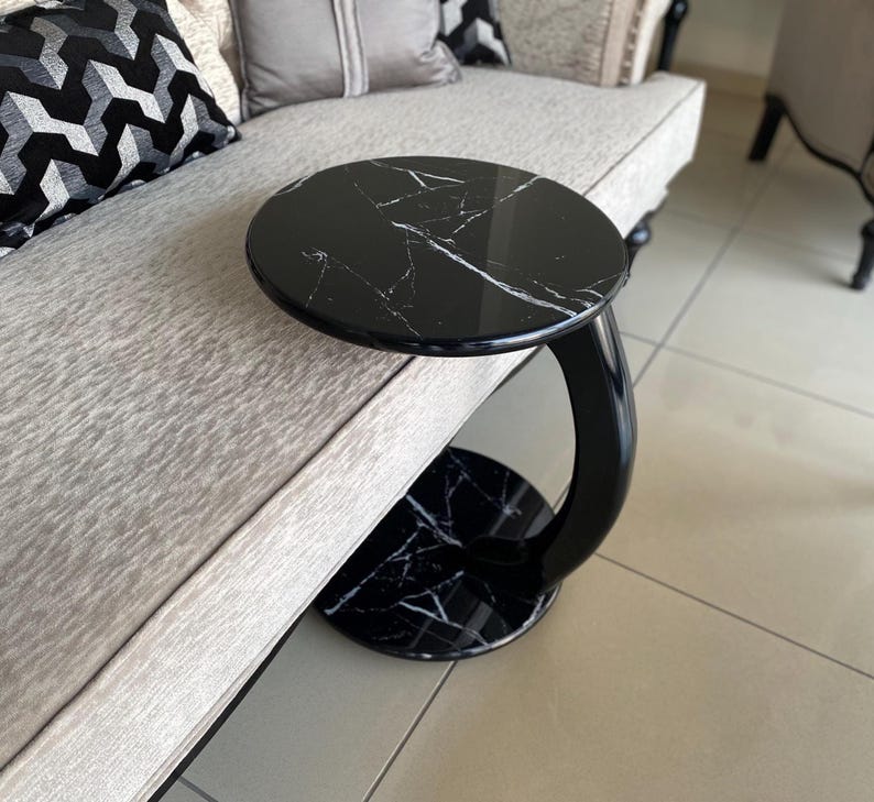 May include: A C-shaped side table featuring a circular black faux marble top and base with white veining. The table has a curved, glossy black support column that connects the top and base, designed to slide over the edge of a sofa or chair.