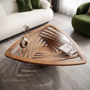 May include: A triangular coffee table with a clear glass top and a base constructed from curved, layered wooden slats. A small, light-coloured ceramic vase and a book titled Design are placed on the glass surface of the table.