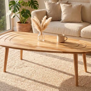 May include: An oval wooden coffee table with four tapered legs and a smooth surface featuring concentric carved grooves. The light-toned wood table holds a clear glass vase containing dried pampas grass and a small ceramic mug on its top surface.