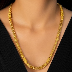 Puede incluir: Un collar de cadena dorada que presenta una serie de eslabones circulares interconectados intercalados con cuentas cilíndricas doradas texturizadas. El collar tiene un acabado pulido y descansa sobre una prenda negra con un patrón repetitivo.