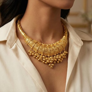 Puede incluir: Un collar llamativo de tono dorado con un diseño de cuello curvo y grueso, patrones en relieve intrincados y un fleco de pequeñas cuentas esféricas colgantes a lo largo del borde inferior. La pieza tiene un acabado metálico pulido.