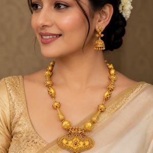 Puede incluir: Un collar de oro con cuentas esféricas texturizadas y un gran colgante ornamentado en forma de corazón con un centro amarillo y detalles en rojo y verde, junto con pendientes jhumka de oro a juego con filigrana intrincada y cuentas.