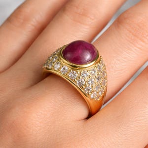 Puede incluir: Un anillo de oro con una gran piedra preciosa de color rojo intenso, tallada en cabujón liso en el centro. La banda ancha está incrustada con varias filas de pequeñas piedras blancas redondas y transparentes que crean un aspecto brillante y reflectante.