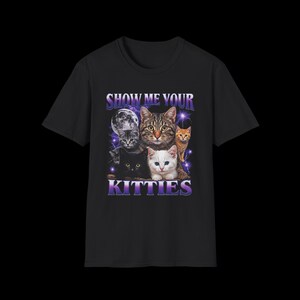 以下が含まれることがあります： 黒の半袖Tシャツで、タビー、黒、白、オレンジなど様々な色の5匹の猫のグラフィックプリントが施されています。デザインには満月、紫色の稲妻、そして紫色の文字でSHOW ME YOUR KITTIESと書かれています。