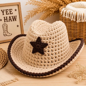 May include: A beige crocheted cowboy hat featuring a dark brown crocheted star centered on the front crown. The hat has a wide, curved brim edged with a dark brown crocheted border. The texture is consistent throughout the crown and brim.