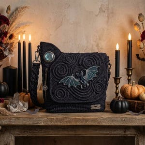 Puede incluir: Un bolso bandolera de ganchillo negro con un patrón de espiral texturizado y un motivo de murciélago bordado en la solapa frontal. El bolso incluye una correa trenzada, un detalle circular iridiscente y una pequeña etiqueta metálica.