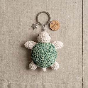 Peut inclure: Un porte-clés tortue en crochet avec une tête et des nageoires couleur crème et une carapace vert nuancé. L'anneau métallique comprend une petite étoile argentée et une étiquette ronde en bois gravée du texte HANDMADE WITH LOVE.