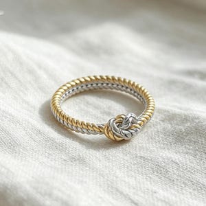 May include: A two-tone ring featuring a twisted rope design in alternating gold and silver-colored metal. The band is formed by two intertwined strands that meet at the front to create a prominent, central knot detail with a textured finish.