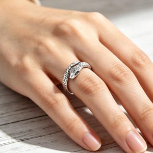 Può includere: Un anello color argento a forma di serpente avvolto, caratterizzato da dettagliate texture a scaglie lungo la fascia e una testa definita con segni oculari scuri. Il metallo presenta una finitura ossidata per evidenziare il motivo.