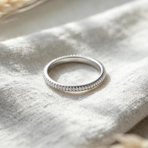 May include: A silver-toned band ring featuring a continuous row of small, clear, sparkling stones set along the outer circumference. The metal has a polished, reflective finish, and the stones are evenly spaced to create a uniform, shimmering appearance.