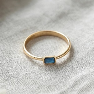 May include: A thin gold band ring featuring a single, small, rectangular-cut blue gemstone set horizontally. The metal has a polished finish, and the stone is securely held in a simple bezel setting, creating a clean and understated appearance.