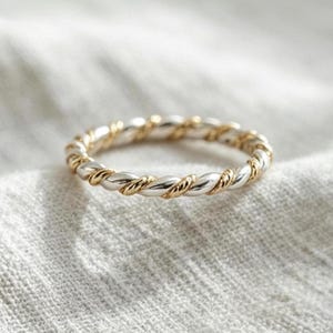 以下が含まれることがあります： 磨かれた銀色の金属と質感のある金色の金属の交互のストランドで構成された、ツイストデザインの細いリング。バンドには、円形のリングの全周を包み込む連続したロープのようなパターンが施されています。