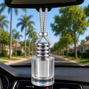 Pode incluir: Um frasco de vidro transparente para ambientador de carro com tampa metálica prateada e um cordão prateado adornado com pequenas contas brancas. O frasco pende do espelho retrovisor, com uma forma cilíndrica e um pescoço prateado estriado.
