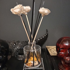 May include: A clear glass reed diffuser bottle containing amber liquid and small decorative stones, featuring three light-colored wooden flower-shaped reeds and several thin black sticks extending upwards from the narrow neck of the container.