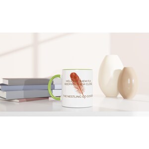 Może przedstawiać: Biały ceramiczny kubek z jasnozielonym uchwytem i wnętrzem. Z przodu znajduje się grafika brązowego pióra z napisem: HELPING THEM FLY, KEEPING THEM CLOSE, THE NESTLING CO GOODS. Kubek jest ustawiony na płaskiej powierzchni.