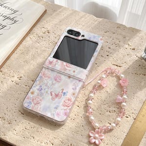 May include: A foldable smartphone case featuring a floral and butterfly pattern in shades of pink, white, and blue, paired with a beaded phone charm made of white pearls, clear pink beads, and a large pink flower charm, next to a ruler.