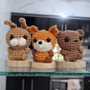 Puede incluir: Tres pequeñas figuras de animales tejidas a ganchillo sobre bases redondas de madera individuales. La figura de la izquierda es un conejo marrón claro, la del centro es un zorro naranja y blanco, y la de la derecha es un oso marrón oscuro.