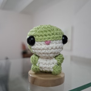 Puede incluir: Una pequeña criatura redonda tejida a ganchillo con hilo verde claro en la parte superior y los lados, y una sección blanca en la cara y el vientre. Tiene dos ojos grandes de plástico negro brillante y una pequeña nariz rosa bordada sobre una base.