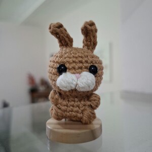 Puede incluir: Una pequeña figura de conejo tejida a ganchillo en color marrón con dos orejas erguidas, ojos de plástico negro y un hocico de hilo blanco con una pequeña nariz rosa bordada. El peluche está montado sobre una base circular de madera clara.