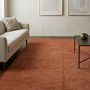 Könnte beinhalten: Ein rechteckiger Teppich in tiefem Terrakotta-Orange mit einer strukturierten Oberfläche, die horizontale erhabene Linien über die gesamte Länge aufweist und durch eine einzelne vertikale Linie in zwei Abschnitte mit parallelen Streifen unterteilt ist.