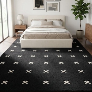 Puede incluir: Una alfombra rectangular con una base de color negro sólido y un patrón repetitivo de pequeños símbolos blancos en forma de cruz, distribuidos uniformemente por toda la superficie. La alfombra tiene una textura de pelo corto.
