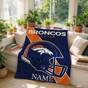 May include: A navy blue and orange fleece blanket featuring the Denver Broncos logo, a football helmet graphic, and the text BRONCOS DENVER NAME. The design includes a large helmet illustration centered on the fabric with bold team colors.