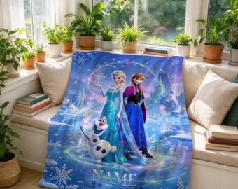 Könnte beinhalten: Eine blaue Fleecedecke mit einem Grafikdruck von Elsa, Anna und Olaf aus Die Eiskönigin. Das Design zeigt wirbelnde Schneeflocken, eine Winterlandschaft und das Wort NAME in weißen Großbuchstaben in der unteren Mitte des Stoffes.