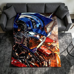 Puede incluir: Una manta rectangular que presenta una ilustración gráfica detallada de un personaje robótico azul y rojo con un casco metálico, ojos brillantes y armadura mecánica, sobre un fondo de color naranja intenso y oscuro.