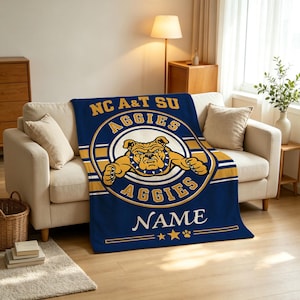 May include: A navy blue fleece throw blanket featuring the NC A&T SU Aggies logo with a central bulldog illustration. The design includes gold and white circular accents, the text AGGIES, a customizable NAME field, and star and paw print icons.