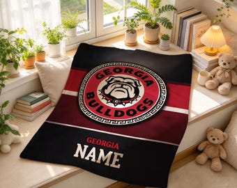 Könnte beinhalten: Eine schwarz-rote Wohndecke mit dem Georgia Bulldogs-Logo in einem kreisförmigen Emblem in der Mitte. Die Decke hat ein horizontales Streifendesign und den Text "GEORGIA NAME" in weißen Großbuchstaben am unteren Rand.