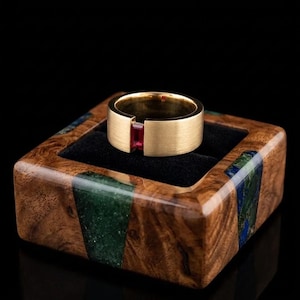 May include: A brushed gold band ring featuring a single rectangular red gemstone set in a tension style. The ring is displayed in a square wooden box with inlaid green and blue stone accents and a black velvet interior lining.