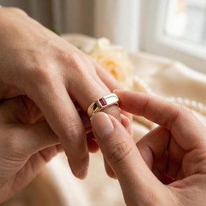 Peut inclure: Une bague en or avec une pierre précieuse rubis rectangulaire taille baguette sertie horizontalement au centre. La bague présente une finition polie et lisse ainsi qu'un anneau en métal solide et large qui s'affine légèrement vers l'arrière.