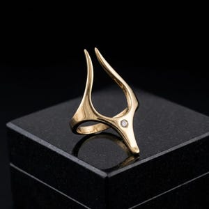 May include: A gold-toned ring featuring an open, elongated V-shaped design that extends upward. The band is smooth and polished, with a single small, clear, round-cut stone set at the base of the V-shape where the two arms meet the main band.