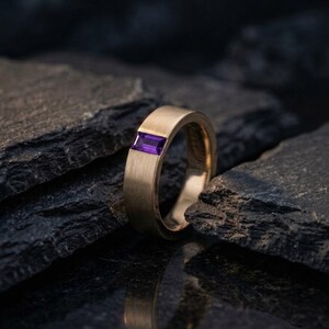 May include: A brushed gold band ring featuring a single rectangular, baguette-cut purple gemstone set horizontally into the metal. The ring has a flat profile and a matte finish, showcasing a clean, geometric design with a vibrant violet stone.