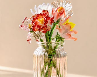 Könnte beinhalten: Eine klare, gerippte Glasvase mit einem Strauß aus handgefertigten Glasblumen. Die Blumen haben zarte Blütenblätter in Rot-, Rosa-, Weiß- und Gelbtönen, wobei grüne Glasstiele durch das transparente, vertikal geriffelte Glas sichtbar sind.