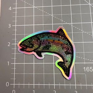 May include: Holographic sticker in the shape of a rainbow trout. The sticker has a black outline and iridescent colors throughout the fish's body. The sticker is on a gray cutting mat with grid lines.