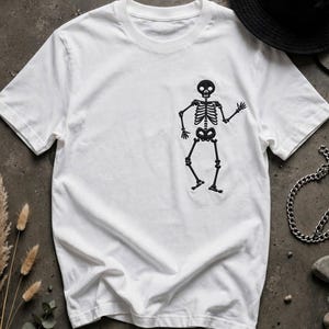 May include: A white short-sleeved t-shirt featuring a black embroidered graphic of a dancing skeleton on the right side of the chest. The skeleton is depicted in a playful pose with arms and legs bent, showcasing detailed ribcage and skull.