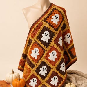May include: A crochet blanket featuring a repeating pattern of square motifs in shades of brown, orange, and yellow. Each square contains a white ghost figure with two black circular eyes. The blanket is draped over a mannequin torso.