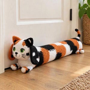 May include: A long, cylindrical crochet door draft stopper shaped like a calico cat. The cat has a white face with green eyes, black whiskers, and long, floppy black and orange ears. Its body features a repeating pattern of orange, black, and white.