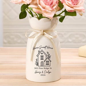 May include: A white ceramic vase with a scalloped rim and a cream-coloured ribbon tied around the neck. The vase features a black graphic of a house with the text Home Sweet Home, 301 Pine Ridge St, Henry & Evelyn, and 2026. Pink roses are inside.