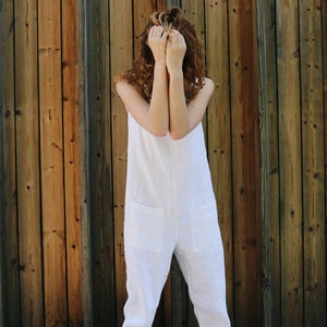 May include: A white sleeveless jumpsuit featuring a round neckline, two large square patch pockets on the front, and tapered pant legs with cuffed hems, paired with brown leather clogs that have a buckle strap and a wooden sole.